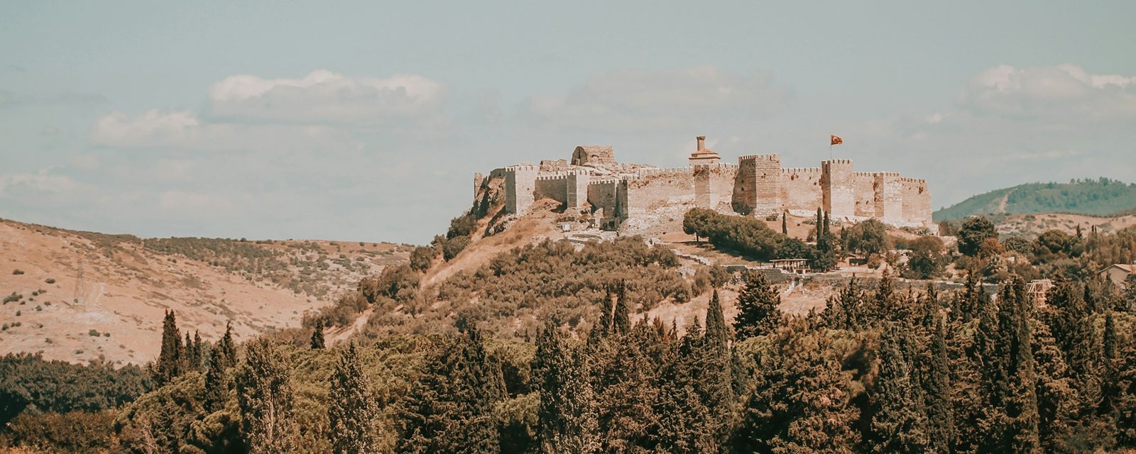 The whole circuit of Ayasuluk Fortress along the hilltop above Selçuk, with square towers, a ruined dome and a minaret stump inside the walls, above slopes of pine and cypress