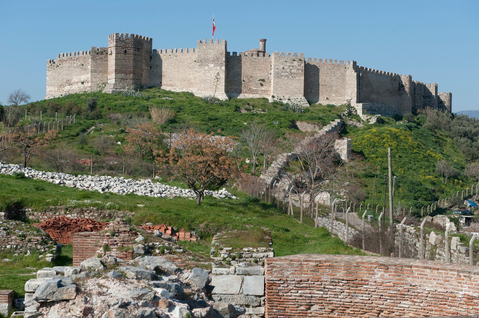 The crenellated walls and square towers of Ayasuluk Fortress running along the green crest of the hill above Selçuk, with a Turkish flag on the walls, seen in spring from the brick ruins of the Basilica of St John