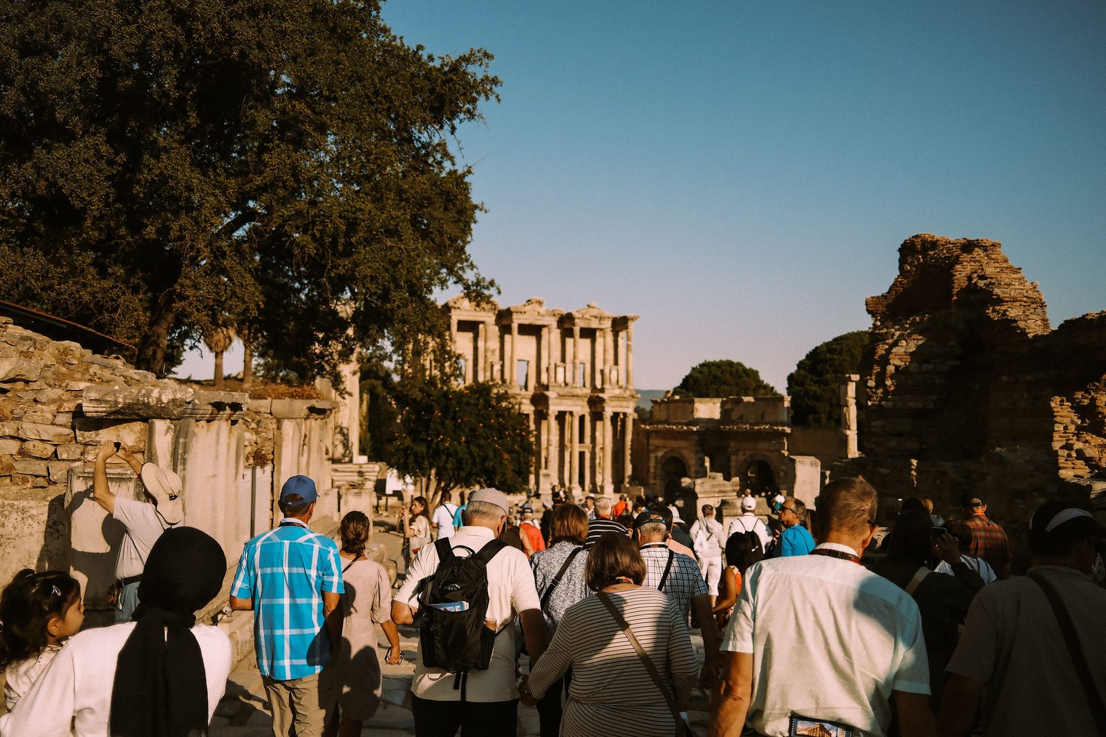 A summer crowd seen from behind on lower Curetes Street in Ephesus, in short sleeves, caps, a sun hat and a headscarf, walking toward the Library of Celsus