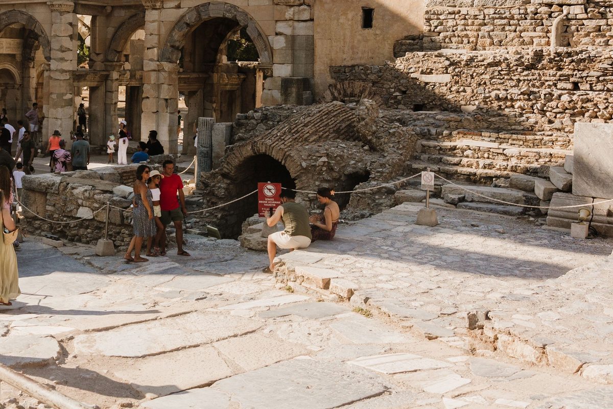 A red sign reading İzinsiz drone uçurmak yasaktır, no drones without permission, on a rope line in front of a vaulted ruin at Ephesus, with visitors nearby and the Gate of Mazaeus and Mithridates behind
