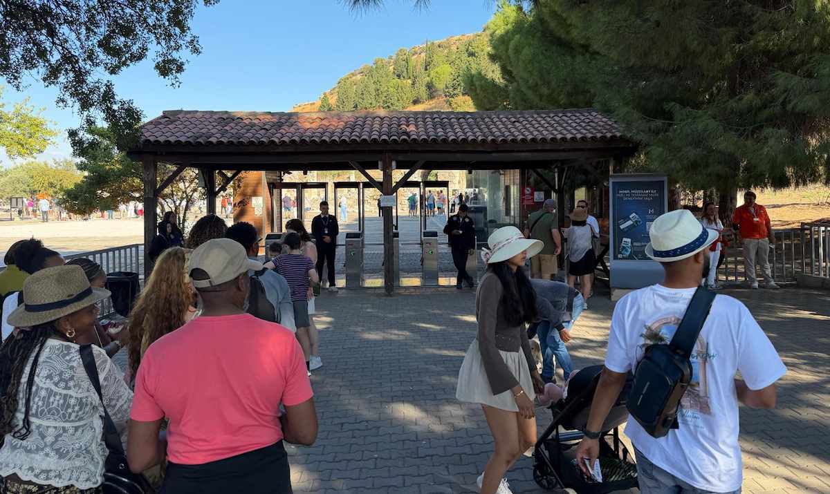 Visitors in sun hats with day bags and a stroller walking toward the turnstiles under the tiled-roof entrance pavilion at the upper gate of Ephesus, with two security staff by the gates and a pine-covered hill behind