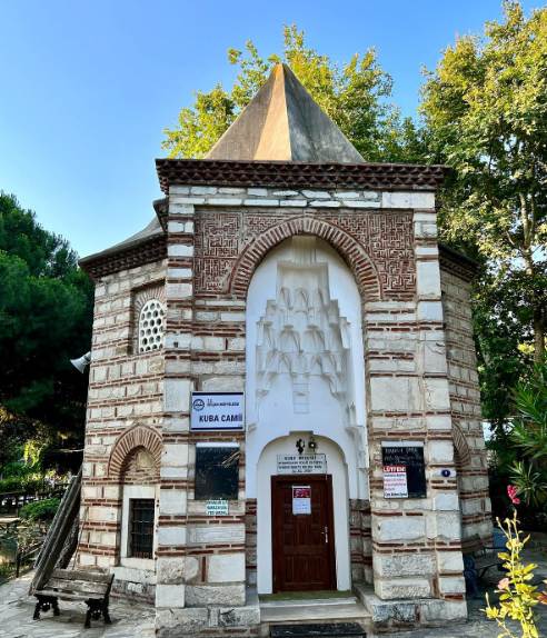 Kuba Camii in Selçuk: a small octagonal mosque built in alternating courses of red brick and pale stone, with a tall projecting portal, a white muqarnas half-vault above a wooden door, panels of geometric brickwork, a gray pyramidal roof and no minaret
