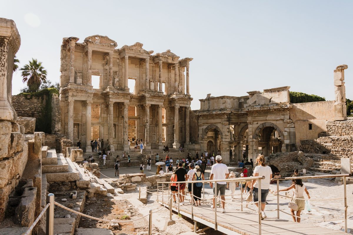 Visitors in summer clothes walking along a rope-lined wooden boardwalk toward the Library of Celsus and the Gate of Mazaeus and Mithridates in Ephesus