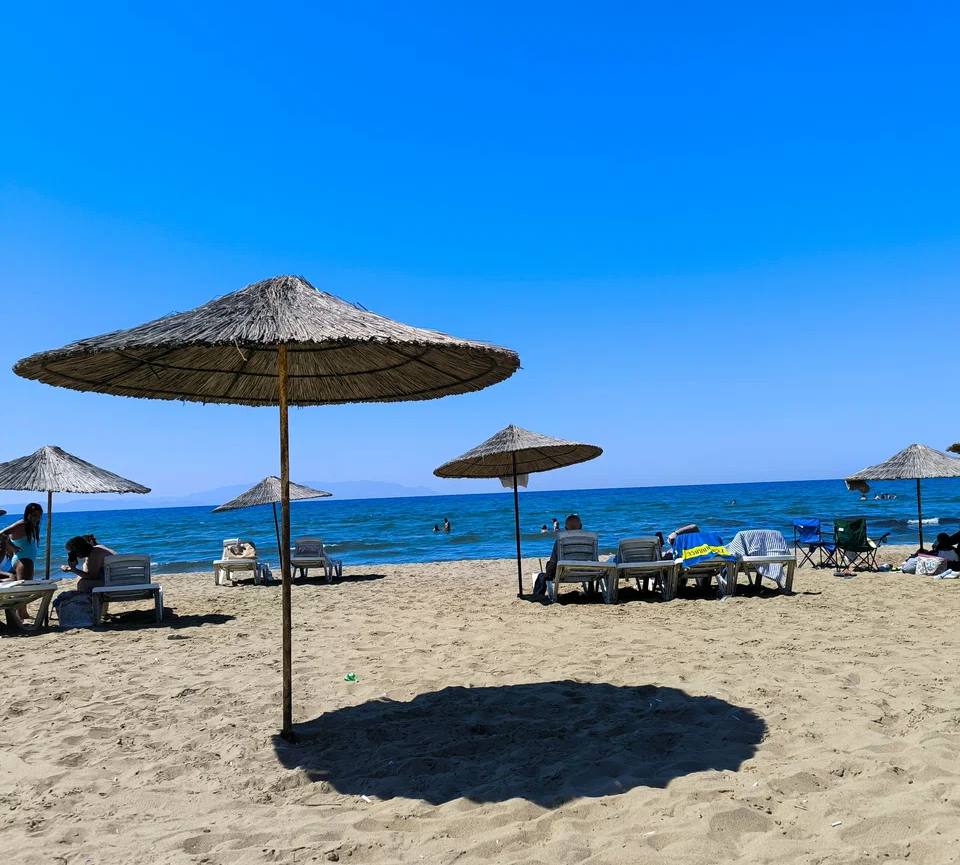 Straw beach umbrellas and white sun loungers on the pale sand of Pamucak beach near Selçuk, with a few swimmers in a calm blue sea