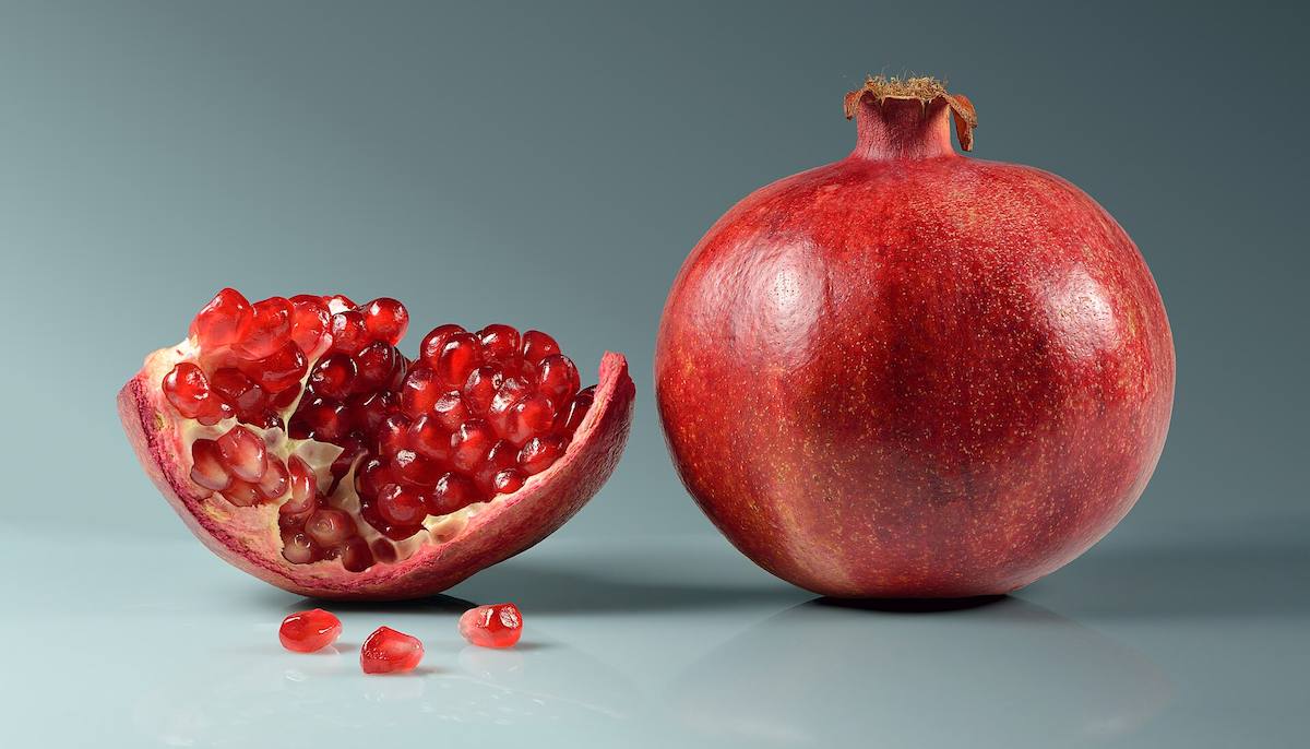 A whole red pomegranate beside an open half packed with glossy red arils, on a plain gray background