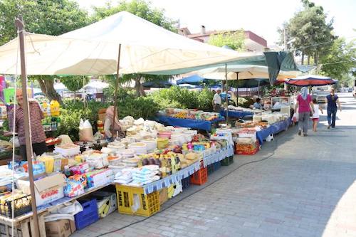 Market stalls under white umbrellas along a tree-lined street in Selçuk, with tubs of cheese and olives, crates and jars on the tables and shoppers walking between them