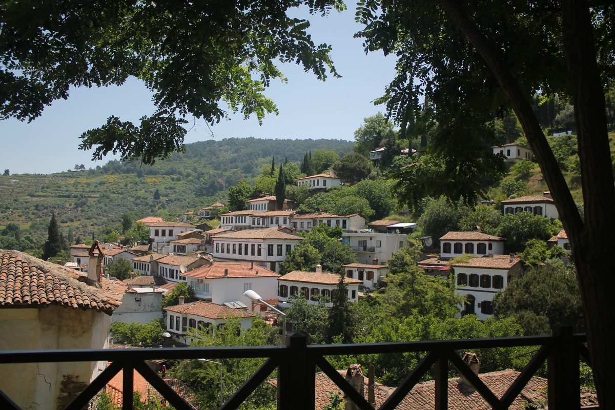White stone houses with tiled roofs spread across a green hillside at Şirince, seen through trees from a wooden terrace
